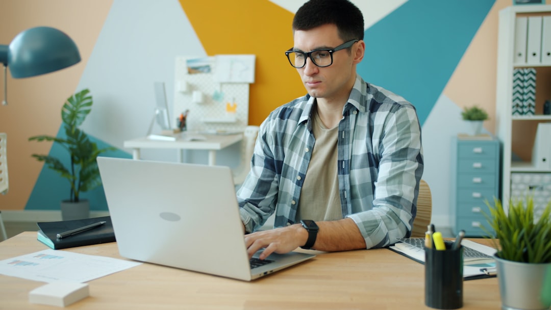 Man with glasses typing on a silver laptop at a wooden desk in a colorful, geometric-office setting
