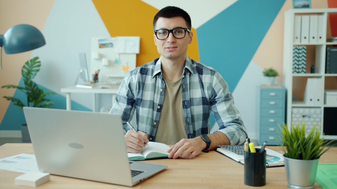 Man with glasses sits at a desk with a silver laptop, writing in a notebook in a colorful geometric office setting.