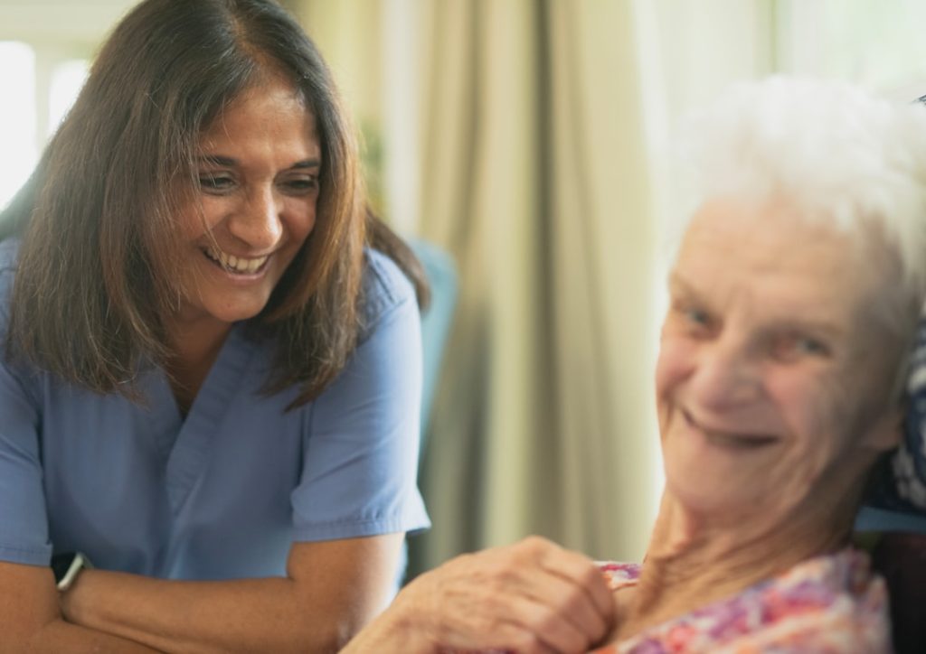 Caregiver in blue scrubs smiling at an elderly woman seated and smiling.