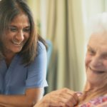 Caregiver in blue scrubs smiling at an elderly woman seated and smiling.
