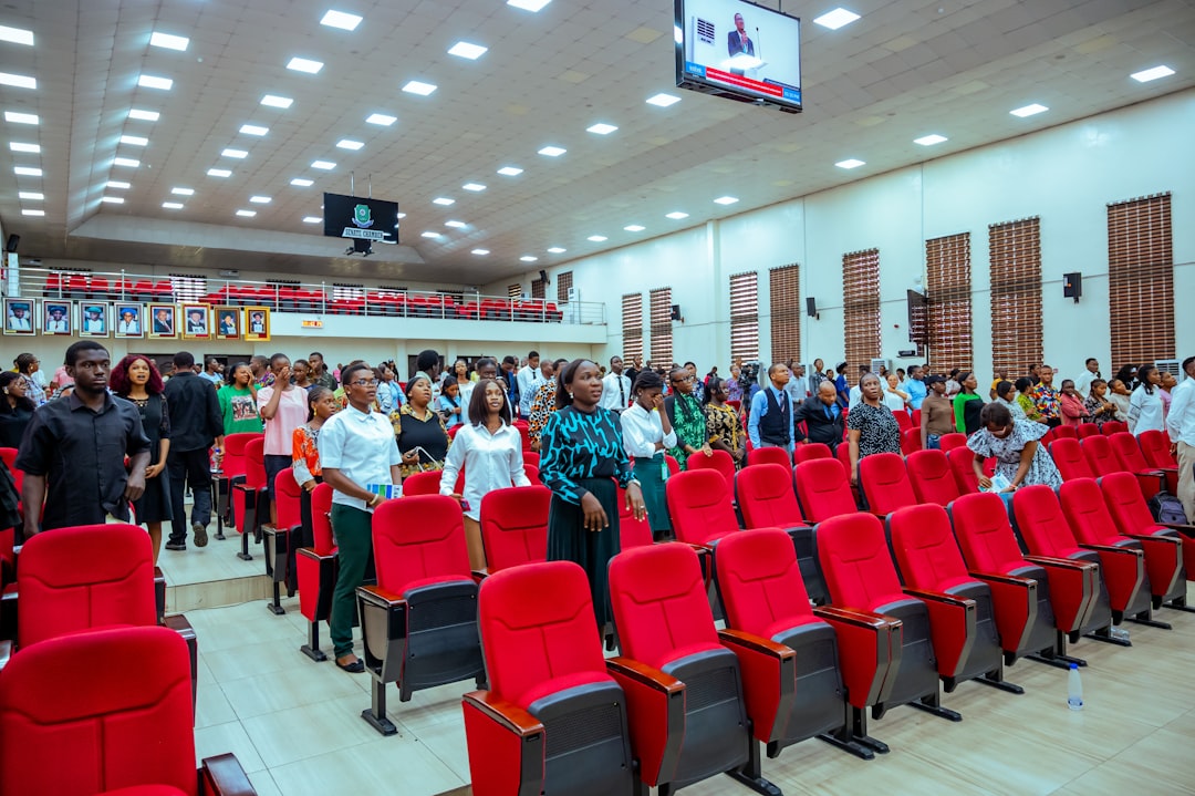 Crowd standing in a modern auditorium with red seats, a large overhead screen, and portraits along the back wall.