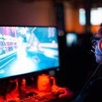 Gamer wearing a headset with glowing red ear cups, intensely focused on a neon-lit computer game on a large monitor.