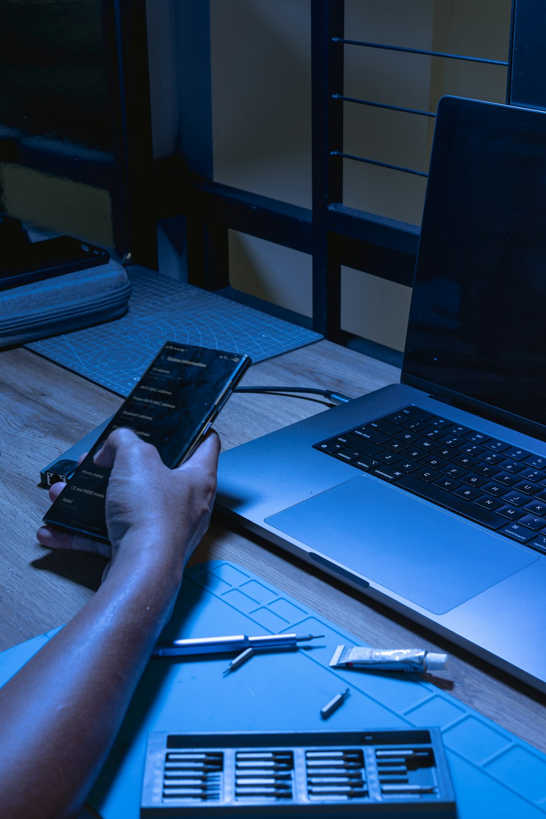 Person's hand holds a smartphone beside a silver laptop on a wooden desk, with a blue cutting mat, pens, and small tools nearby under cool blue lighting.