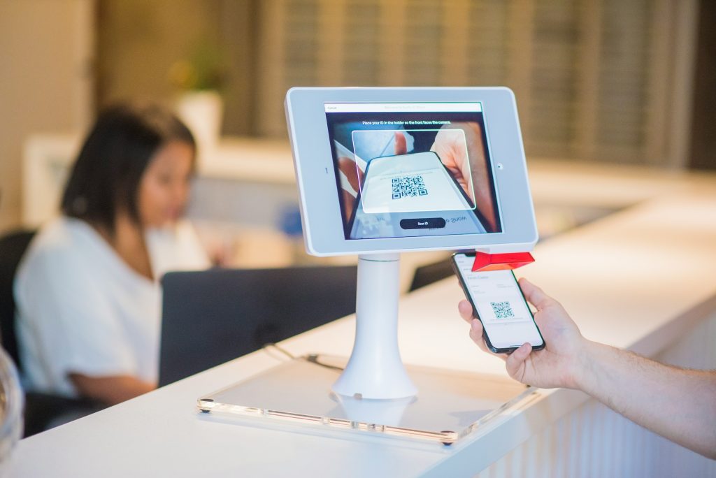 Customer presents a phone QR code to a self-service scanner at a counter, tablet screen showing the QR code being read