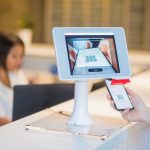Customer presents a phone QR code to a self-service scanner at a counter, tablet screen showing the QR code being read