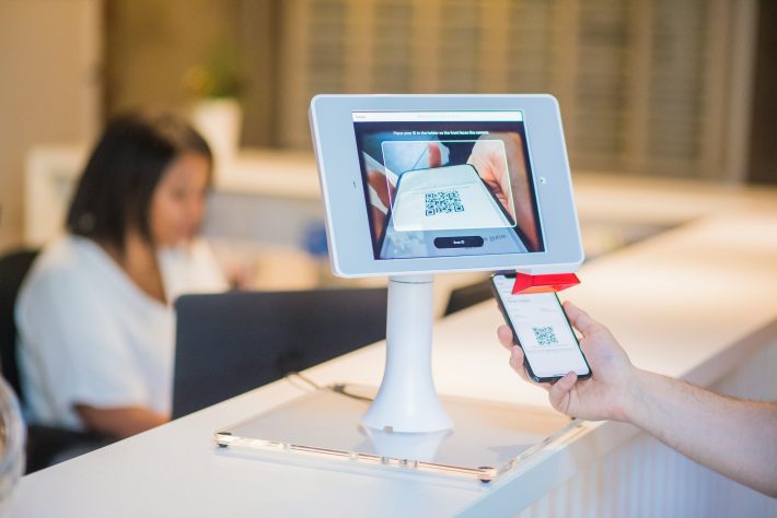 Customer presents a phone QR code to a self-service scanner at a counter, tablet screen showing the QR code being read