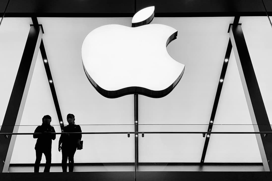 Inside a sleek Apple Store, a large illuminated Apple logo dominates a glass wall; two silhouetted people stand on a balcony beneath.