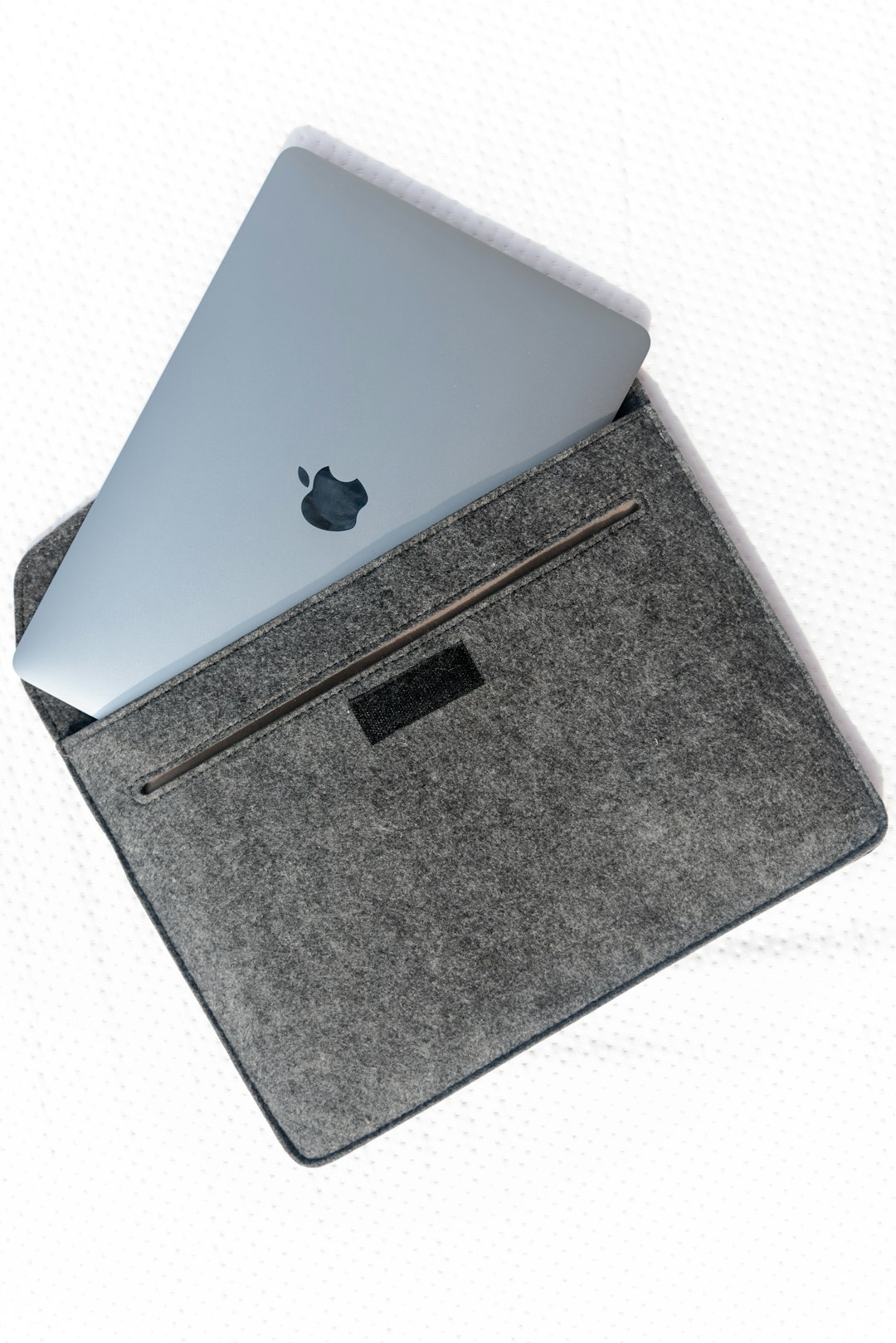 Light blue-gray Apple MacBook partially tucked into a dark gray felt laptop sleeve on a white textured surface.