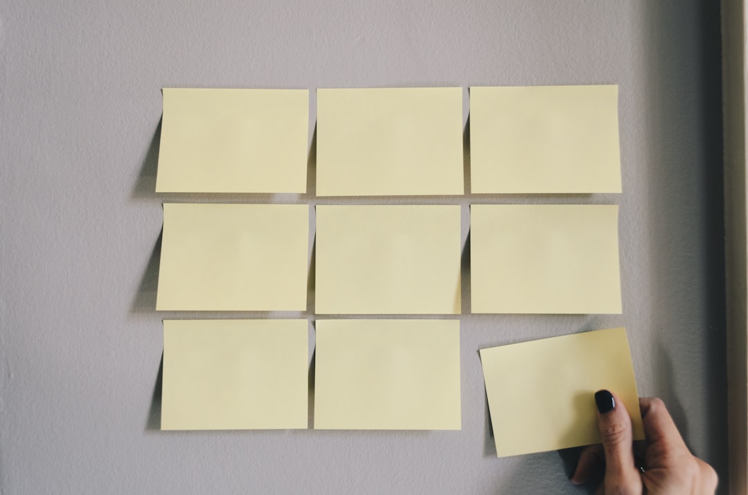 Nine yellow sticky notes arranged in a 3×3 grid on a wall, with a hand placing the bottom-right note.