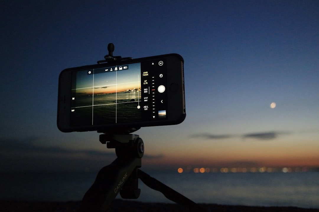Phone mounted on a tripod/gimbal filming a sunset over the water with city lights in the distance and a visible moon.