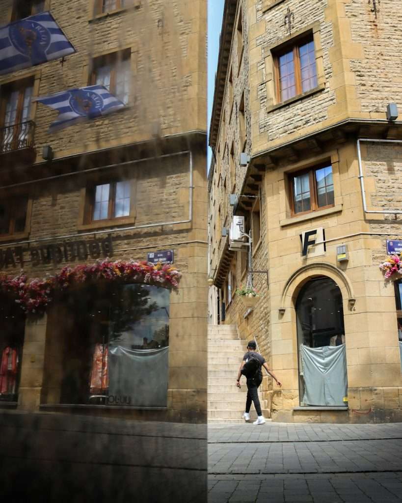 A man walks along a cobblestone street between two stone buildings with arched doorway and stairs, storefronts and flags above.