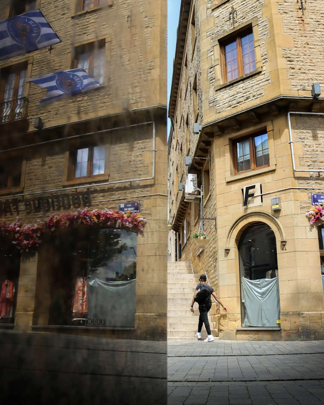 A man walks along a cobblestone street between two stone buildings with arched doorway and stairs, storefronts and flags above.