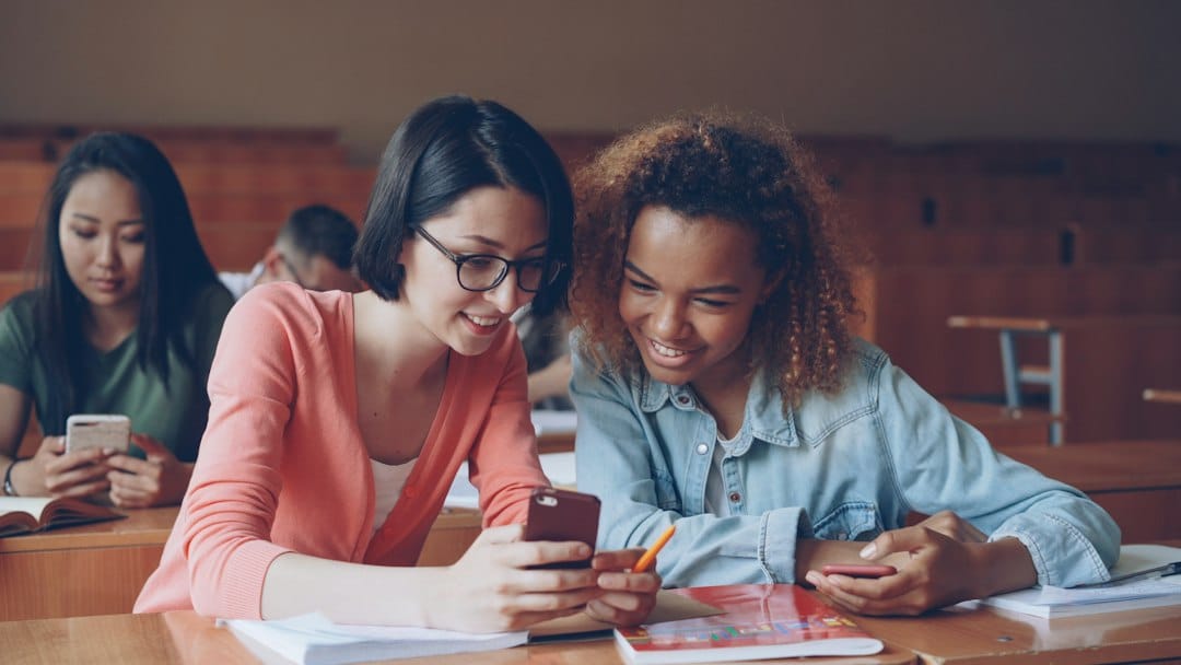 Two students sit at a classroom desk, smiling as they look at a smartphone together.