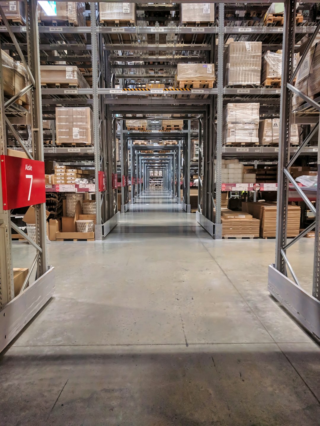 Long warehouse aisle with tall metal shelves stocked with pallets and boxes; a red Aisle 7 sign is visible on the left.