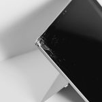 Black-and-white close-up of a smartphone with a cracked screen resting on a slim stand on a light background.