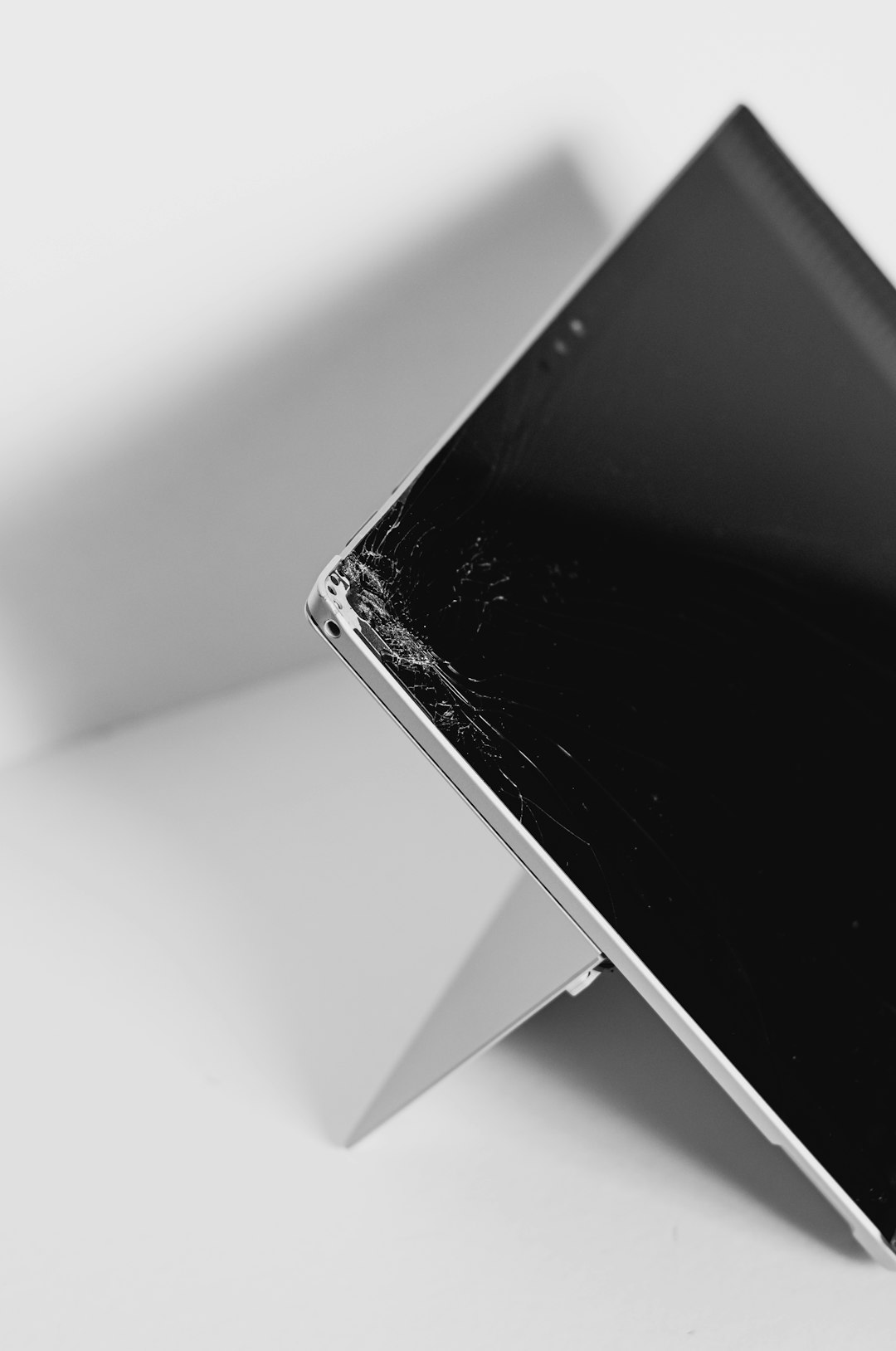 Black-and-white close-up of a smartphone with a cracked screen resting on a slim stand on a light background.