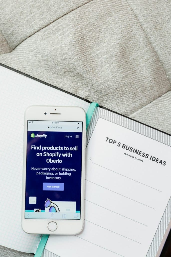 White iPhone displaying the Shopify page beside a notebook titled 'Top 5 Business Ideas' on a gray couch cushion/sofa.