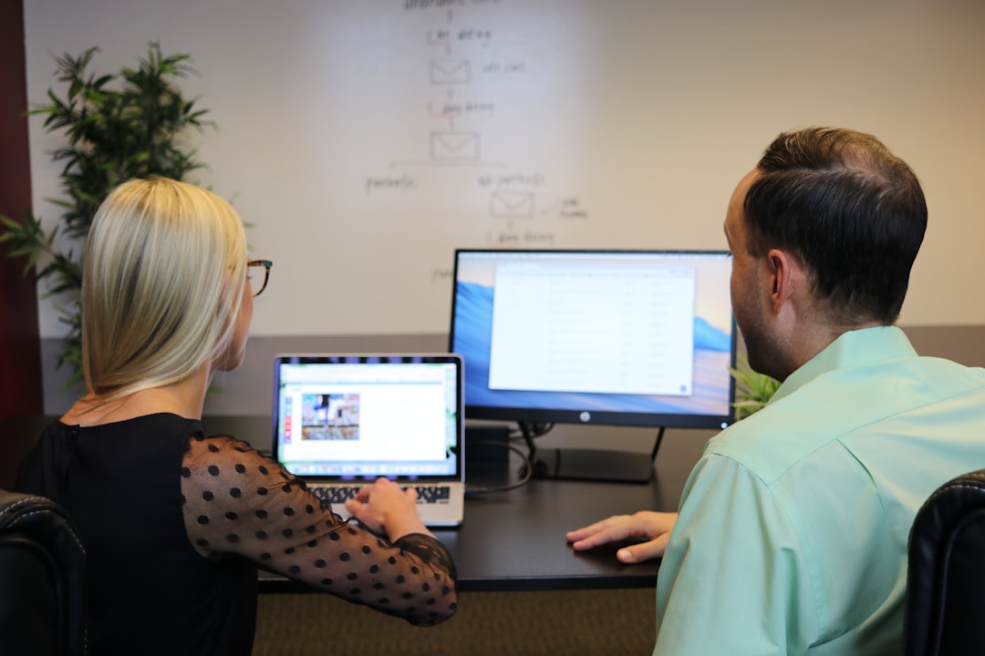 Two coworkers collaborate at a desk, looking at a laptop and a large monitor in a modern office.