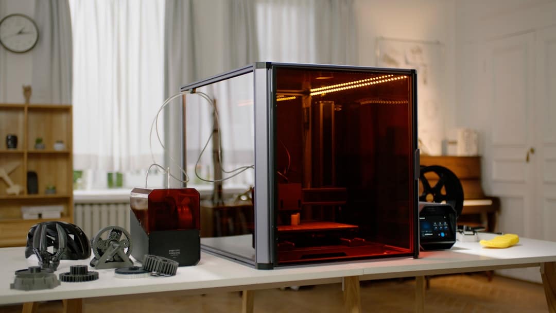 3D printer with red-tinted enclosure on a worktable, surrounded by gears and printer parts in a workshop
