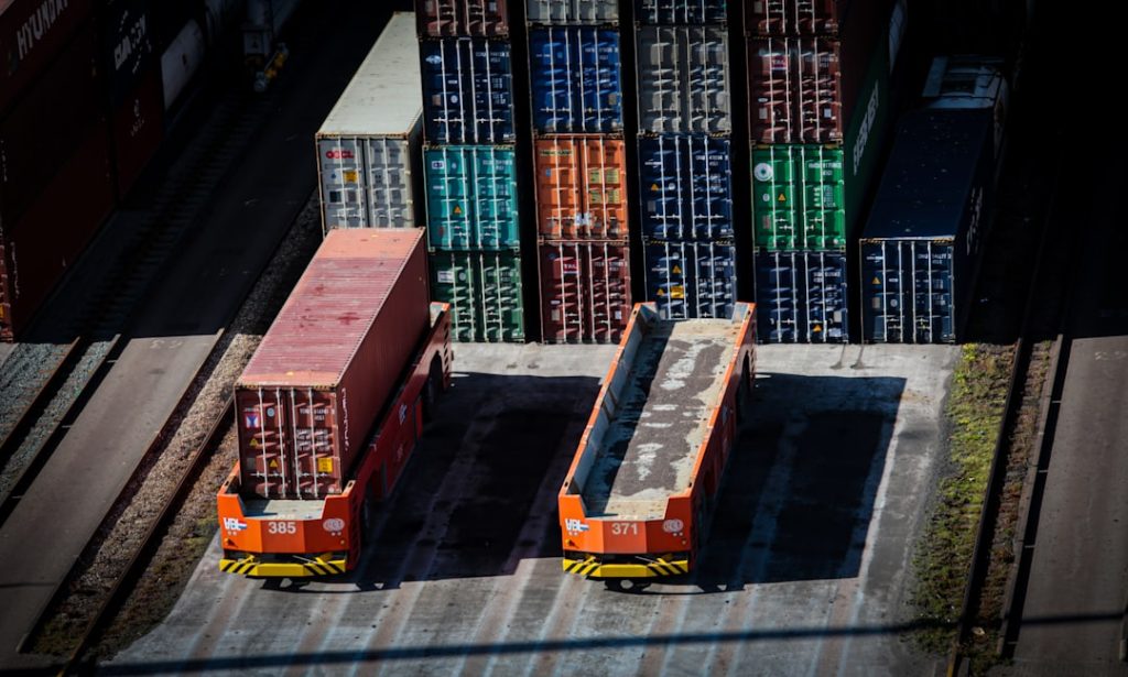 Two orange terminal tractors sit side by side in a container yard with stacked colorful shipping crates behind them.
