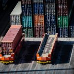 Two orange terminal tractors sit side by side in a container yard with stacked colorful shipping crates behind them.