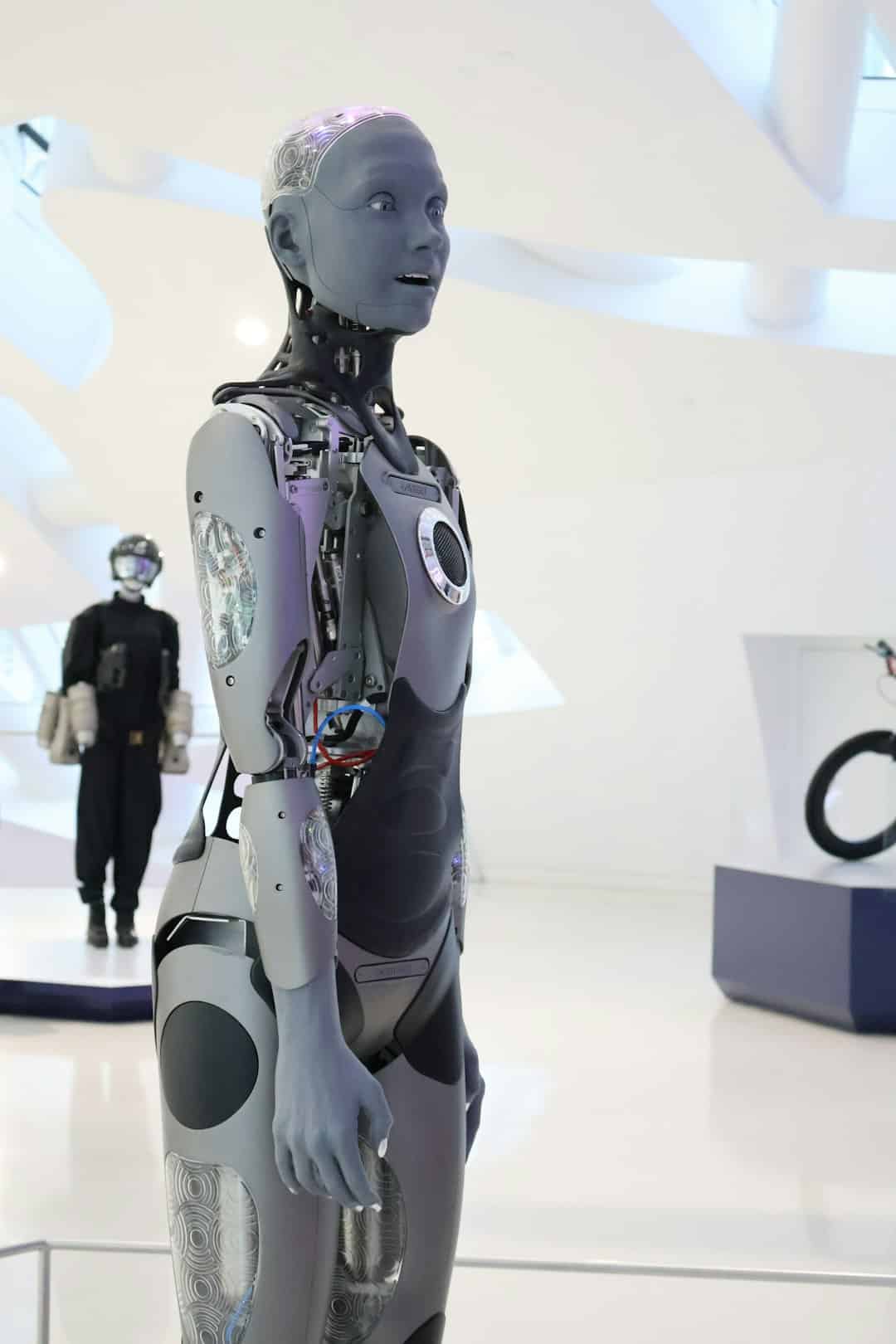 Life-size humanoid robot on display in a bright white gallery; exposed circuitry and purple-gray panels.