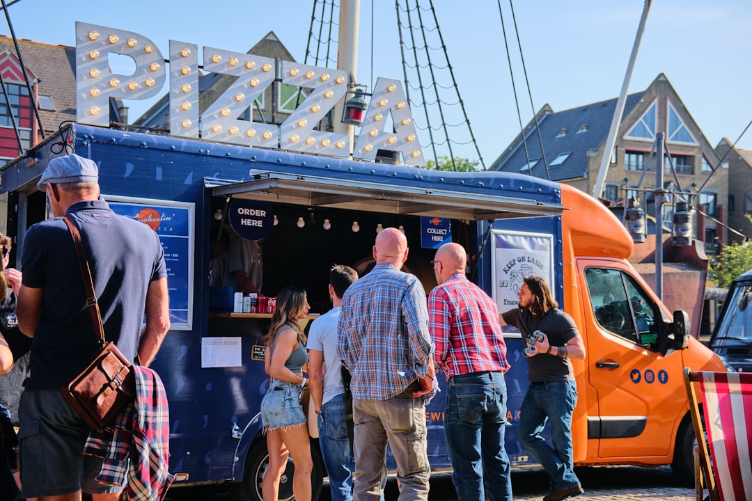 Blue pizza truck with a large illuminated 'PIZZA' sign, people lining up to order on a sunny day, storefronts in the background.