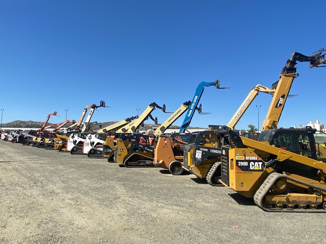 Line of construction skid-steer loaders with raised booms parked on a gravel lot under a clear blue sky, extending in a row.