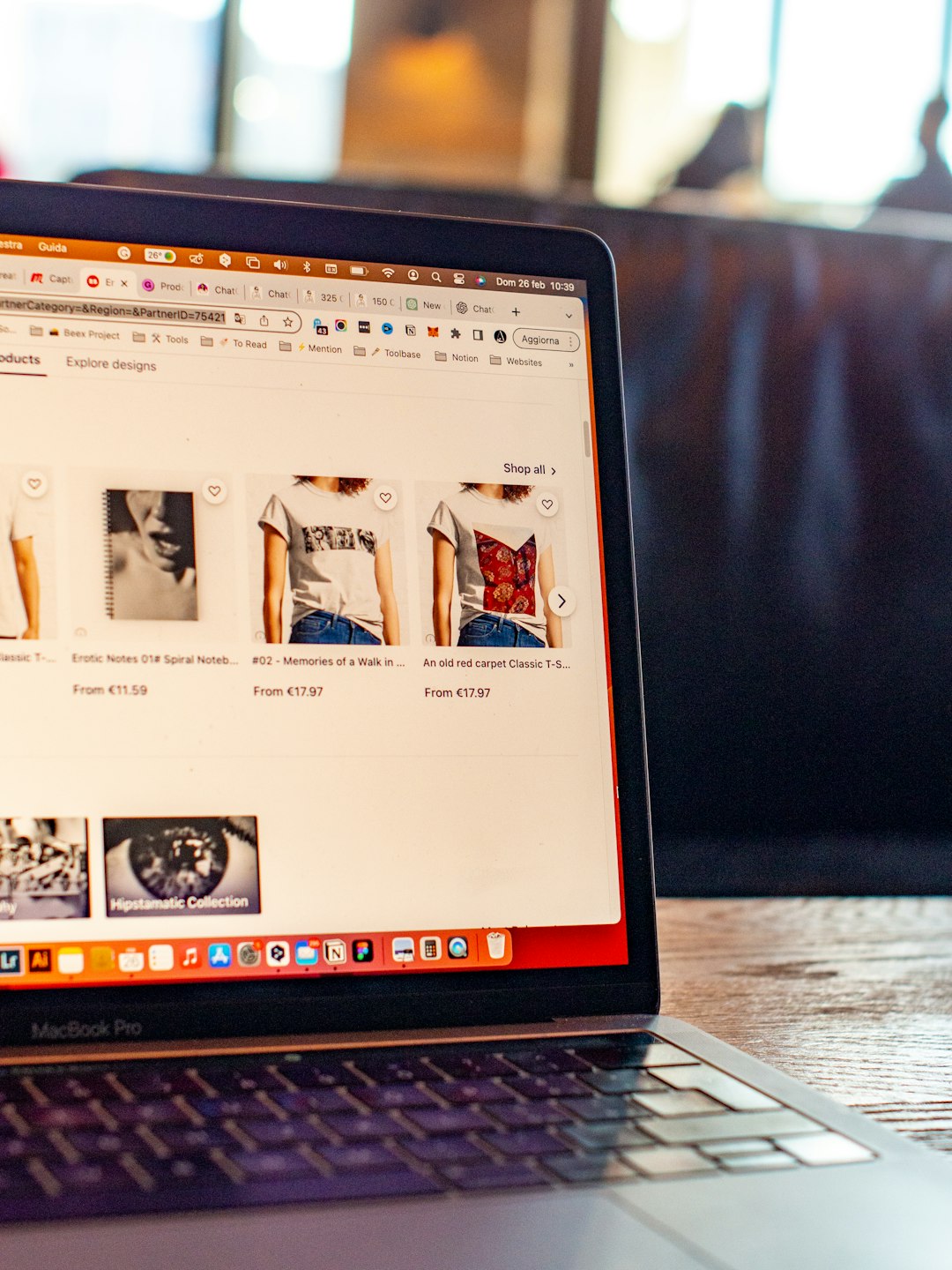 Laptop open to an online fashion store showing t‑shirts on a grid with prices underneath the images.