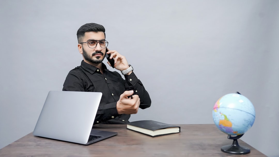 Man wearing glasses and a black shirt sits at a wooden desk, talking on a cell phone with a laptop and a notebook nearby and a globe on the right.