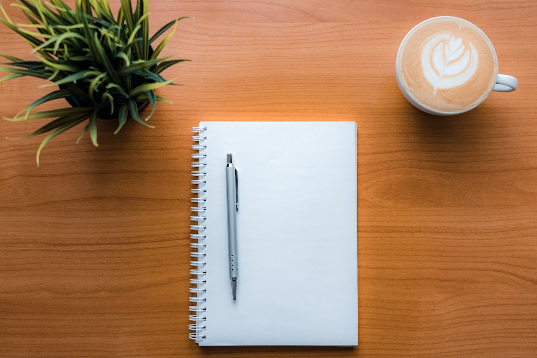 White spiral notebook with a silver pen on a wooden desk, flanked by a potted plant and a latte cup with art on the right.