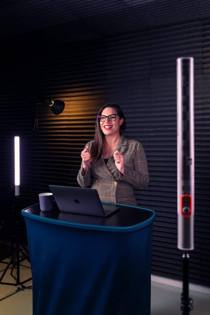 Women presenter at a blue podium with laptop, speaking in a recording studio with lighting.