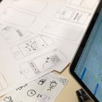 Desk with hand-drawn smartphone wireframes on paper and a tablet showing a map UI beside them, suggesting app prototyping.