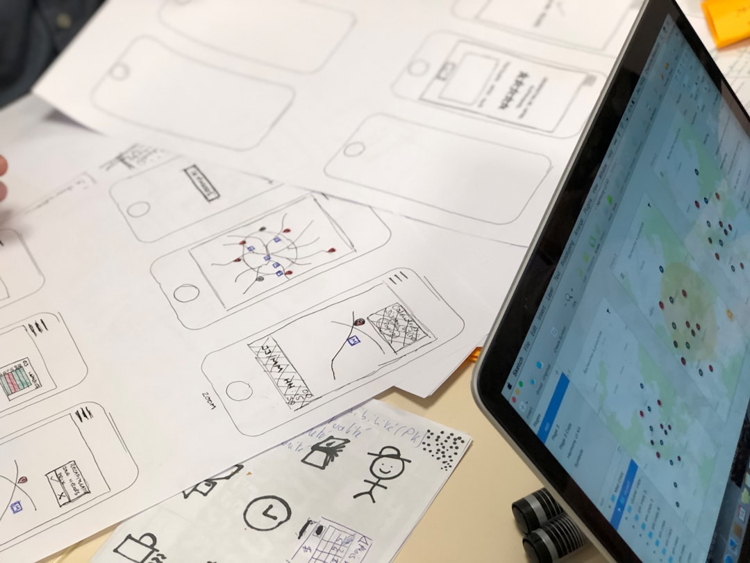 Desk with hand-drawn smartphone wireframes on paper and a tablet showing a map UI beside them, suggesting app prototyping.