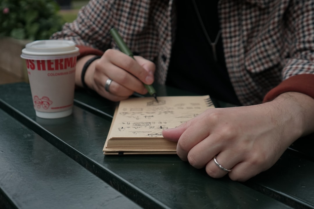 Person writing in a small notebook with a green pen at a green table, a takeaway coffee cup beside them.
