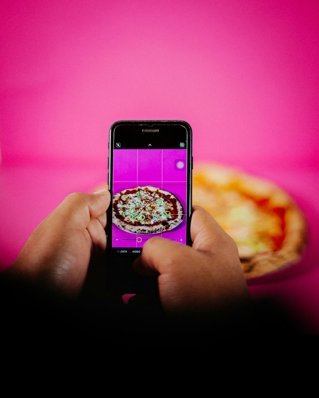 Hands holding a smartphone and photographing a pizza against a bright pink background.