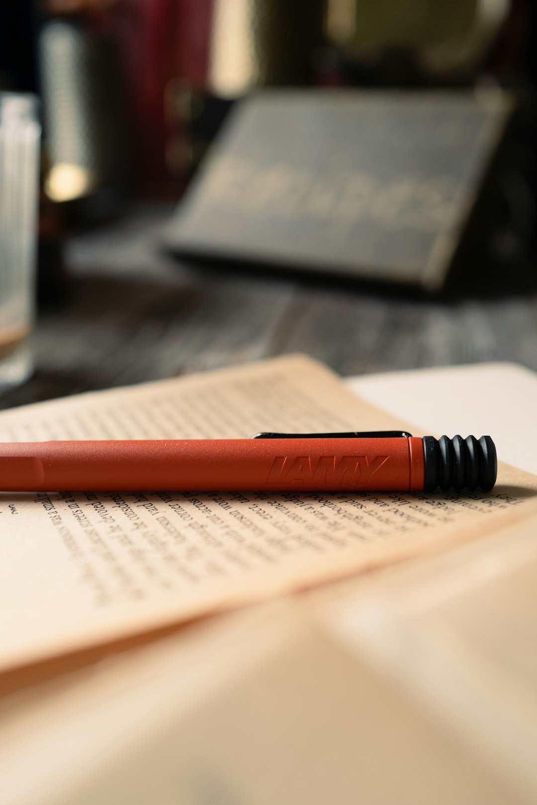 Red LAMY pen resting on an open book with small print, blurred tablet in the background, desk scene.