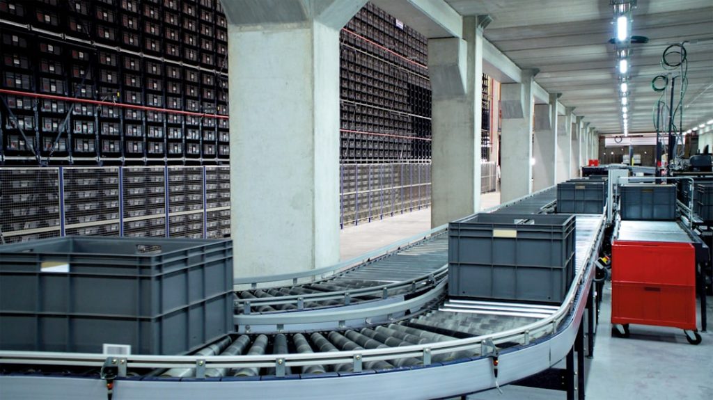 Industrial warehouse with a curved conveyor belt and gray storage bins under bright overhead lighting.