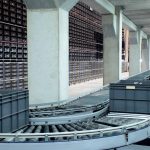 Industrial warehouse with a curved conveyor belt and gray storage bins under bright overhead lighting.