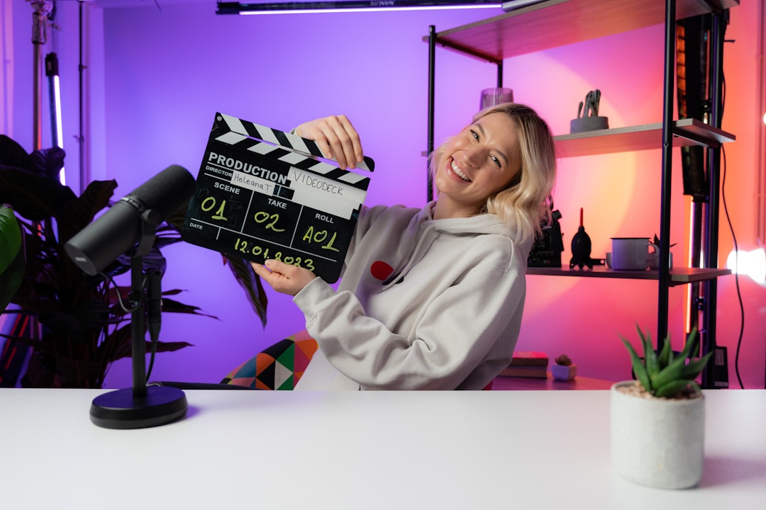 Smiling woman in a hoodie holds a clapperboard on a colorfully lit studio set with a microphone in the foreground.