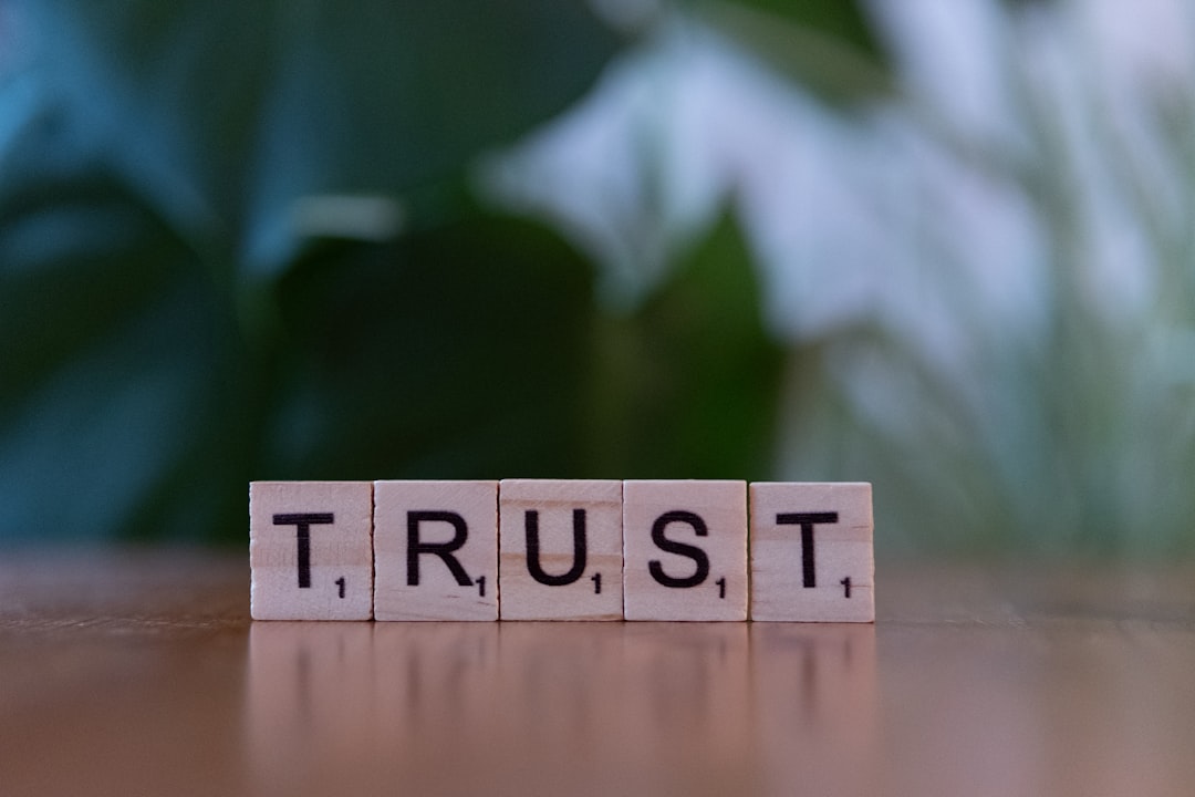 Wooden letter blocks spell “TRUST” on a wooden table with a blurred green background nearby.