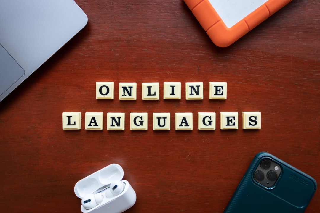 Scrabble tiles spell 'ONLINE LANGUAGES' on a wooden desk beside a laptop, earbuds case, tablet, and a smartphone, suggesting online language learning.