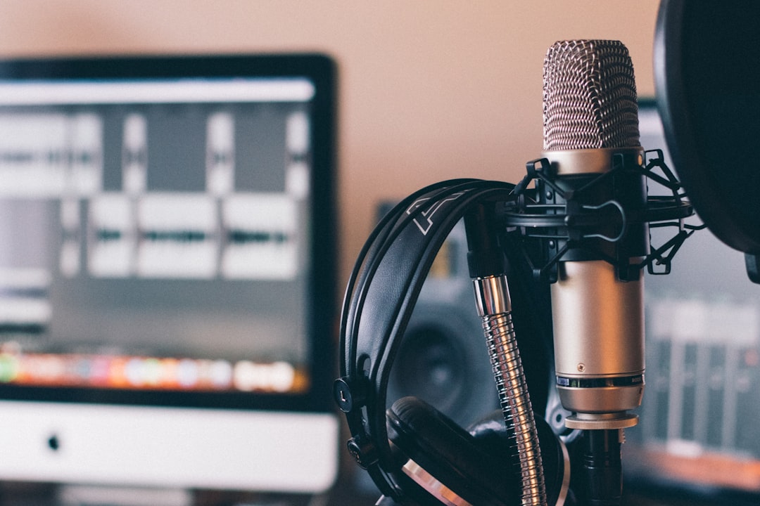 Close-up of a studio microphone with a shock mount and headphones in front of a blurred computer screen in a recording setup