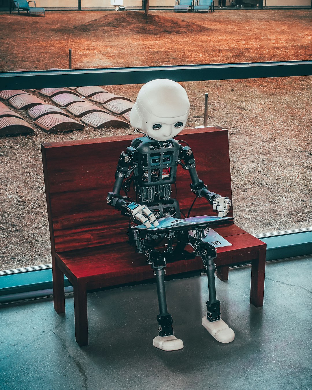 Humanoid robot with a white head and exposed circuitry sits on a wooden bench indoors, looking at a tablet.