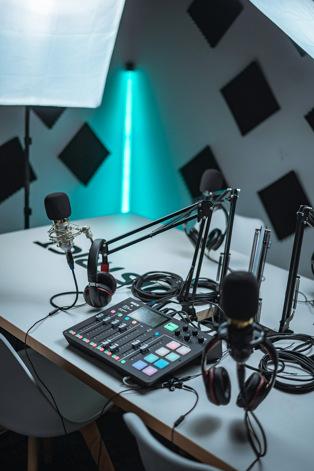 Podcast recording setup with two boom-arm mics, mixer, and closed-back headphones on a table in a studio with teal lighting and acoustic panels.