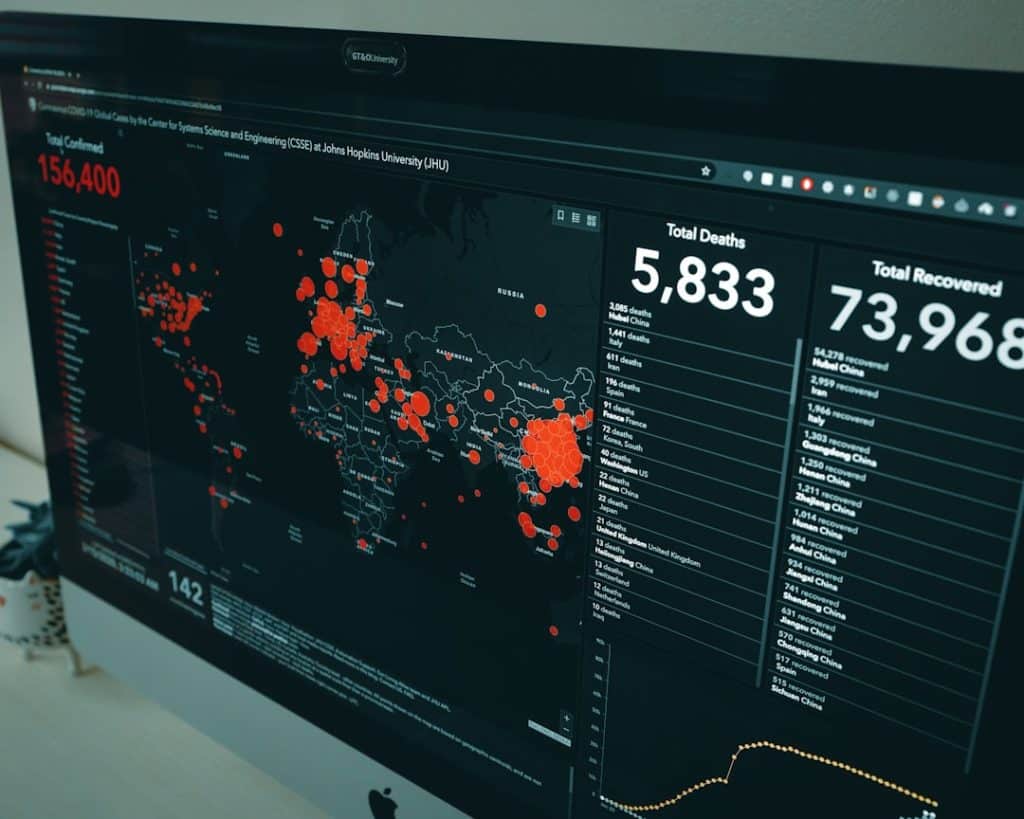 Dark dashboard on a monitor showing a world map with red outbreak markers and right-side stats for deaths and recoveries (e.g., 5,833 deaths; 73,968 recovered).