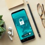 Smartphone on a desk displaying a security app with a padlock and “Secured” badge on a teal world-map background, beside a plant and glasses.