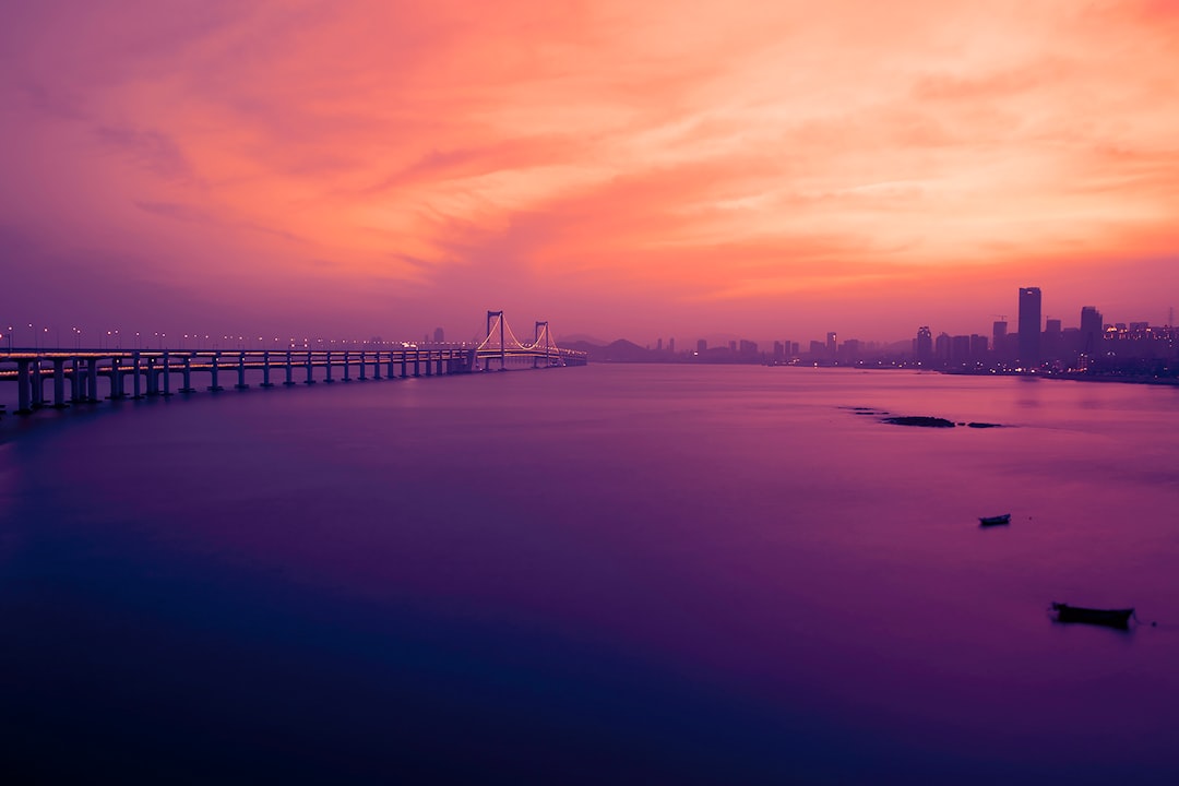 Sunset over a city with a long bridge extending into the water, bathed in pink and orange hues, calm harbor in the foreground.