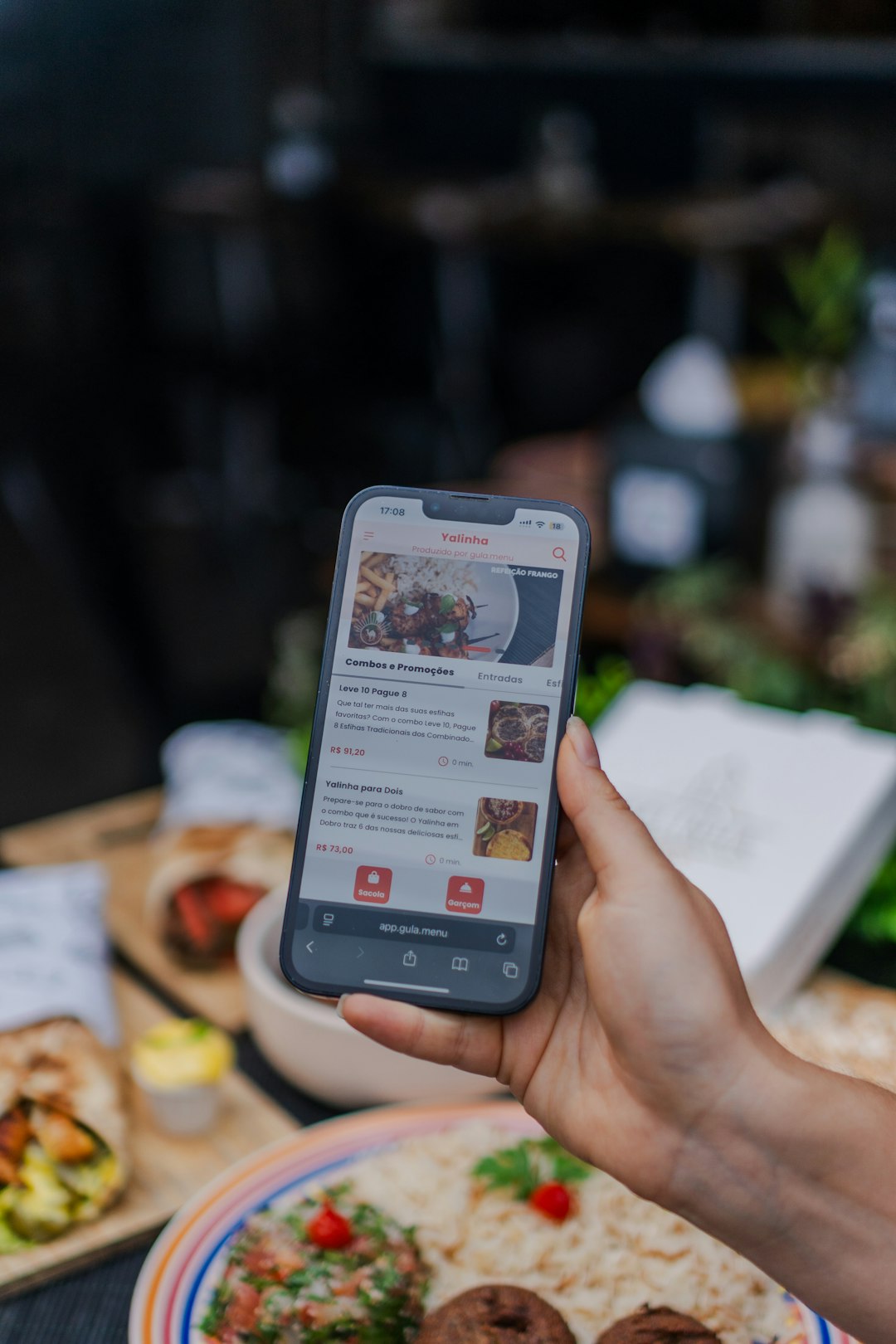 Hand holding a smartphone showing a food delivery app with menu items on a dining table behind it