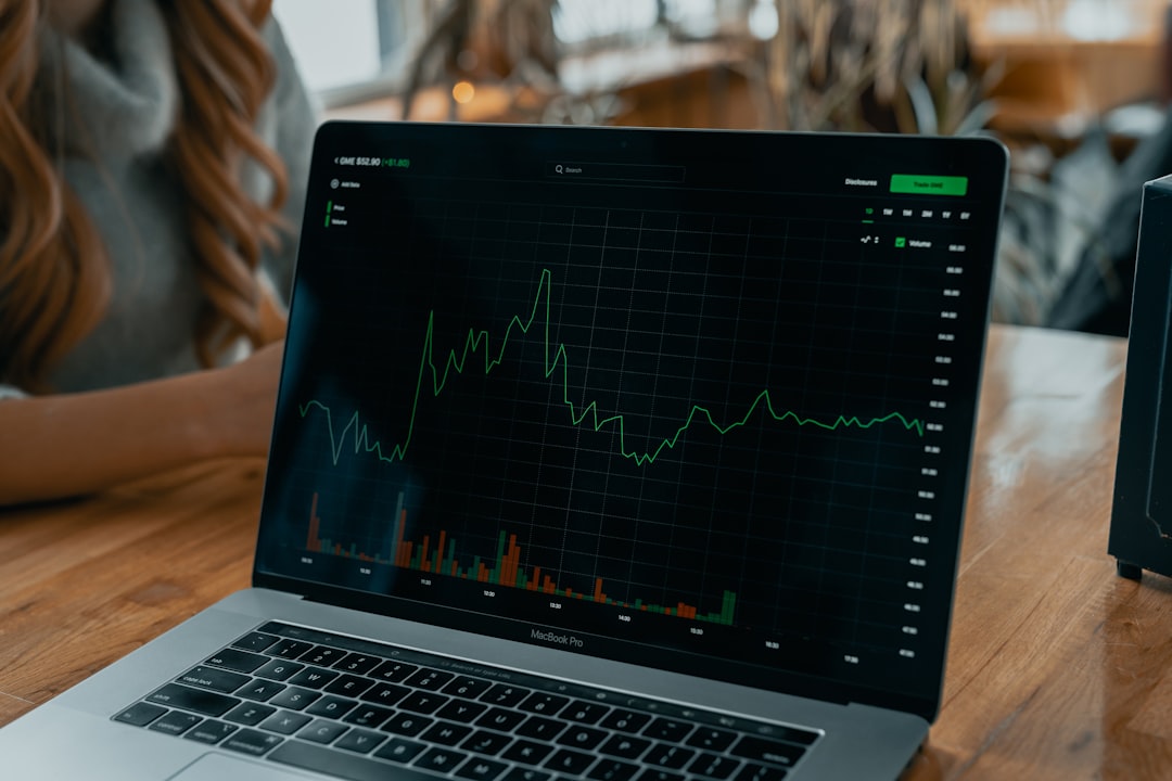 Laptop on a wooden desk displaying a financial chart with a green price line and color-coded volume bars on a dark grid background.